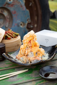 A dish featuring a pile of sushi rolls topped with shredded crab and creamy orange sauce, served on a black ceramic plate. Nearby, there is a bamboo steamer with pickled vegetables, a set of chopsticks, and a small dish of soy sauce. The rustic background includes a blue and rust-colored metal wheel, enhancing the outdoor dining setup.