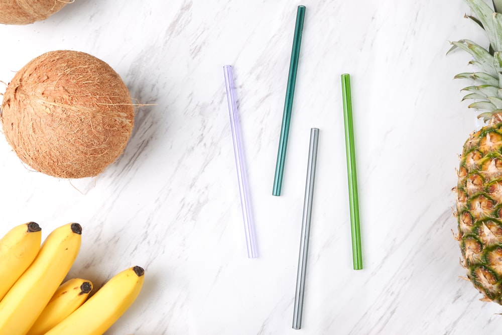 four assorted-color straws beside coconut, bananas, and pineapple