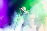 A performer is singing energetically on stage surrounded by vibrant colored lights and thick smoke. The atmosphere is lively and immersive, with the lights creating a dynamic backdrop. Another person appears to be playing a guitar, adding to the musical ambiance.