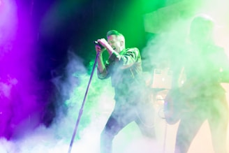 A performer is singing energetically on stage surrounded by vibrant colored lights and thick smoke. The atmosphere is lively and immersive, with the lights creating a dynamic backdrop. Another person appears to be playing a guitar, adding to the musical ambiance.