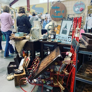 A busy marketplace or flea market stall displaying various decorative items including custom signs, ornaments, and art pieces. The stall is cluttered with wooden tables, small wooden signs, a red metal decoration, and a mix of rustic and modern decor. People are browsing and examining the items, with some individuals standing and looking around. A small pumpkin and other ornaments are placed strategically around the setup.