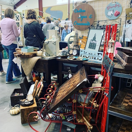 A busy marketplace or flea market stall displaying various decorative items including custom signs, ornaments, and art pieces. The stall is cluttered with wooden tables, small wooden signs, a red metal decoration, and a mix of rustic and modern decor. People are browsing and examining the items, with some individuals standing and looking around. A small pumpkin and other ornaments are placed strategically around the setup.