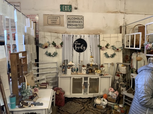 A vintage-themed interior setup with a variety of decorative items displayed on a cabinet. Floral wreaths and small ornamental pieces, such as candles and vases, adorn the area. Signs above the display read 'Leashed Dogs Only' and 'Alcoholic Beverages Prohibited.' The scene includes hanging decorations, a small table with books and plants, and a person wearing a blue jacket.