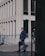 A man in a blue suit walks down a city street, holding a black briefcase. The background features a modern building with large windows and a railing. Another person is partially visible, walking in the opposite direction.