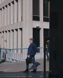 A man in a blue suit walks down a city street, holding a black briefcase. The background features a modern building with large windows and a railing. Another person is partially visible, walking in the opposite direction.