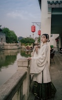A person in traditional attire stands by a riverside railing, holding a fan. The background features a serene river, lush greenery, and traditional architecture with red lanterns hung overhead.
