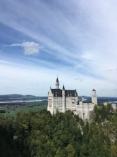 A majestic castle with multiple spires sits atop a lush, green hill. Vast fields and a serene body of water extend in the background under a partly cloudy sky.