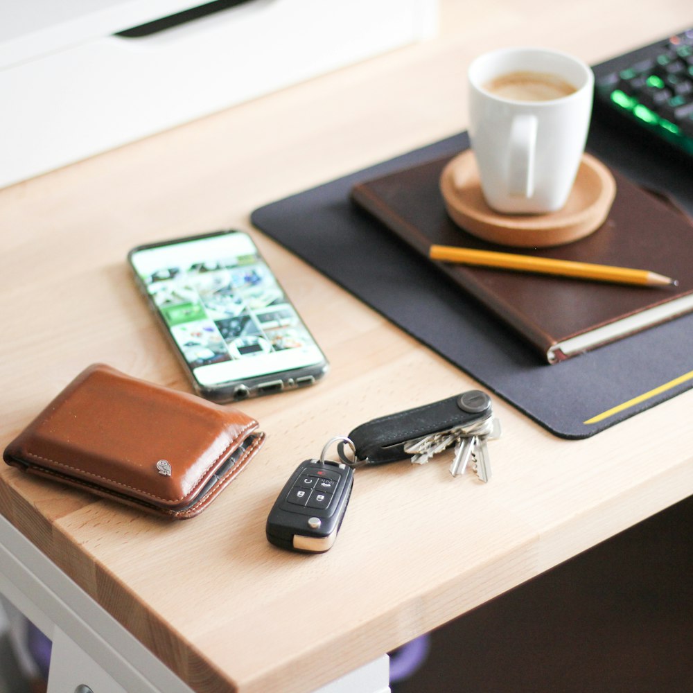 yellow wooden pencil on brown hardbound book near cappuccino, silver Android smartphone, brown leather bifold wallet, and black car fob