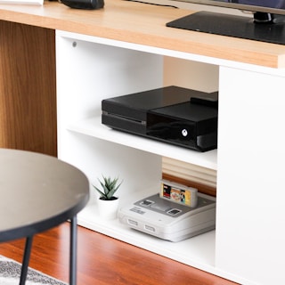 A black gaming console is situated on the middle shelf of a modern, white wooden entertainment unit. Below it, an older version of a gaming console sits with a game cartridge inserted. A small potted plant adds a touch of greenery next to the vintage console. The top of the unit is made of a light wood, and a flat-screen monitor is partly visible on it. The setup is minimalistic and clean, with a round black table in the foreground.