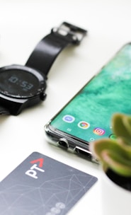 black Android smartphone near round black smartwatch, car fob, leather watch, and green succulent plant