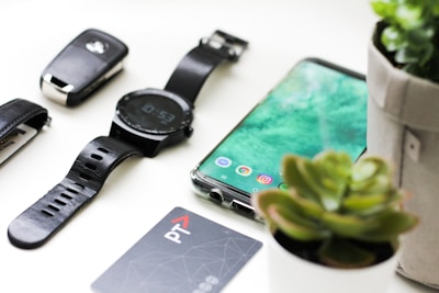 black Android smartphone near round black smartwatch, car fob, leather watch, and green succulent plant