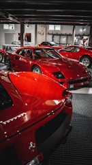 A collection of bright red sports cars is displayed in an indoor showroom with industrial elements. The cars are classic and performance models, positioned closely together. There are people visible in the background, possibly inspecting the vehicles.
