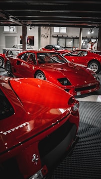 A collection of bright red sports cars is displayed in an indoor showroom with industrial elements. The cars are classic and performance models, positioned closely together. There are people visible in the background, possibly inspecting the vehicles.