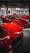 A collection of bright red sports cars is displayed in an indoor showroom with industrial elements. The cars are classic and performance models, positioned closely together. There are people visible in the background, possibly inspecting the vehicles.