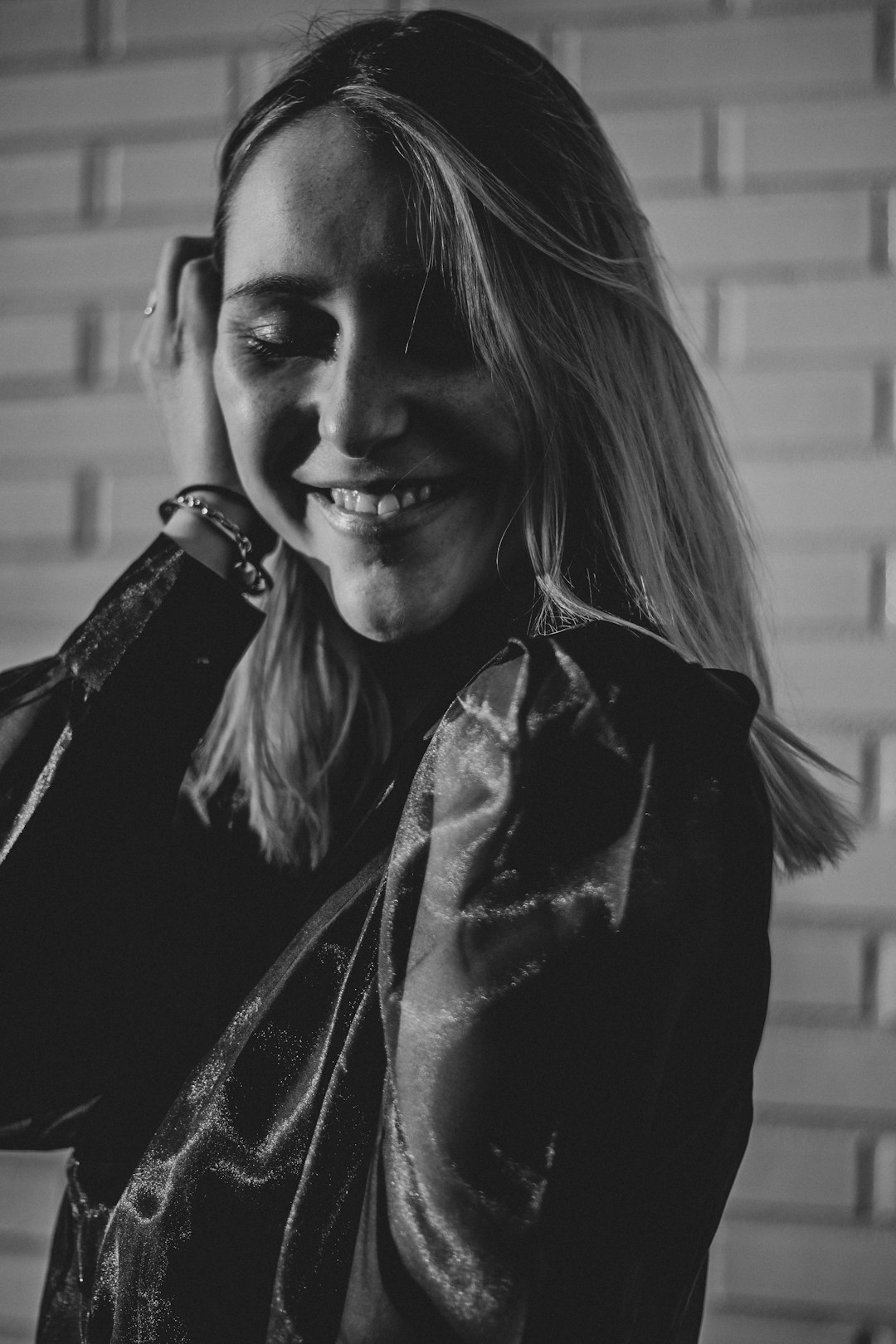 grayscale photography of smiling woman wearing long-sleeved shirt standing while touching her hair