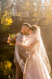 A couple embraces in a tender and intimate moment, surrounded by soft golden lighting. The bride, wearing a flowing veil and delicate gown, holds a bouquet of flowers. They stand near a serene body of water with the backdrop of sunlit autumn trees.