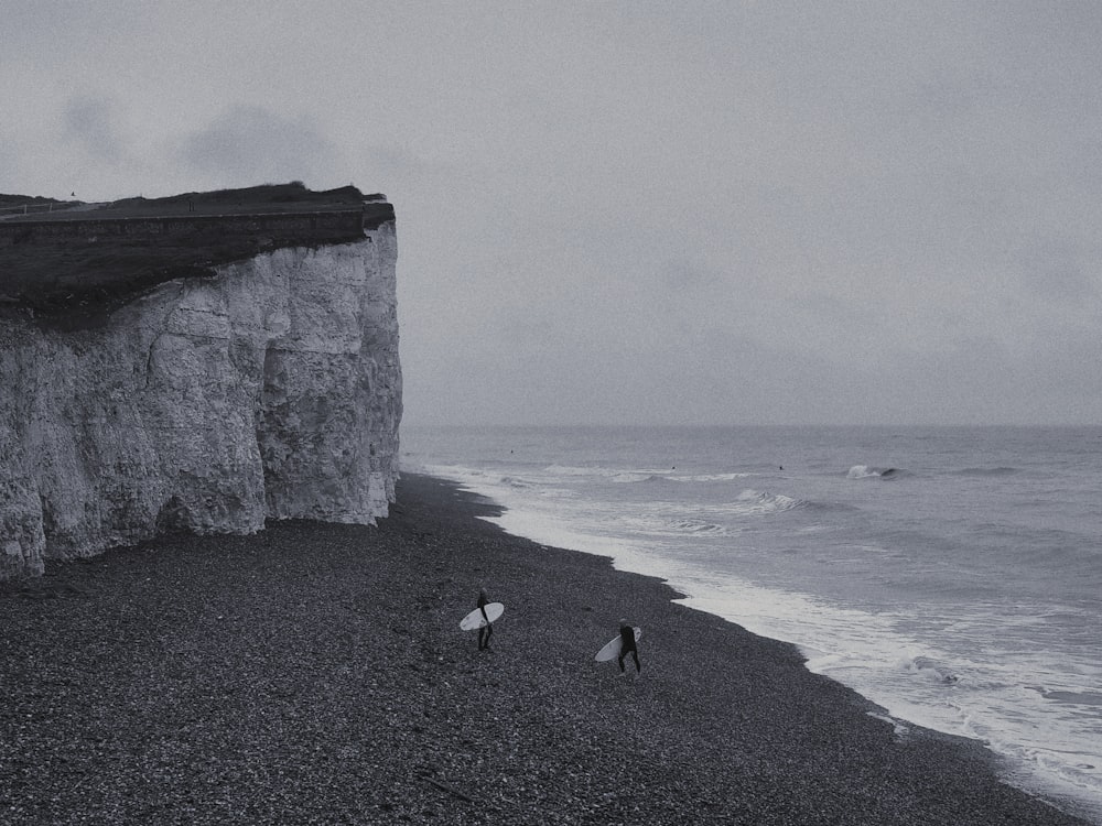 grayscale photography of two person holding surfboard walking near cliff and sea