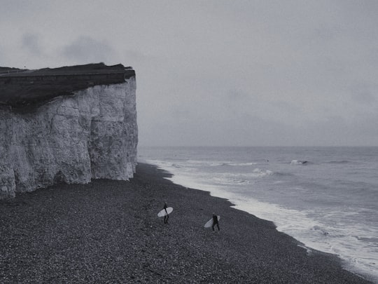 grayscale photography of two person holding surfboard walking near cliff and sea in Seven Sisters United Kingdom