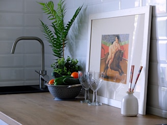 A kitchen countertop with a modern sink, framed artwork, plants, glassware, and a bowl of fresh vegetables. The artwork depicts a person riding a bull. The arrangement creates a harmonious blend of art and culinary elements.
