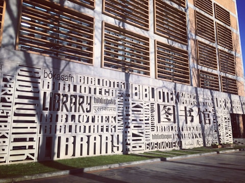 A modern building facade prominently features the word 'library' in multiple languages, artistically integrated into the design. The structure has a grid-like pattern of windows and shutters.