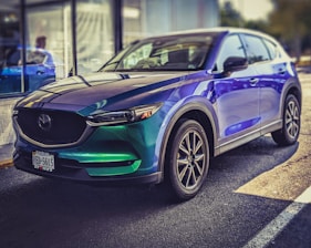 A shiny, modern SUV is parked on a sunny day. The car features a vibrant color gradient with shades of green and blue, complemented by sleek, black-rimmed wheels. Reflections of light create patterns on its surface, enhancing the car's contemporary design. The background includes a windowed building and hints of greenery, suggesting an outdoor environment.