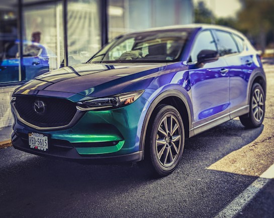 A shiny, modern SUV is parked on a sunny day. The car features a vibrant color gradient with shades of green and blue, complemented by sleek, black-rimmed wheels. Reflections of light create patterns on its surface, enhancing the car's contemporary design. The background includes a windowed building and hints of greenery, suggesting an outdoor environment.