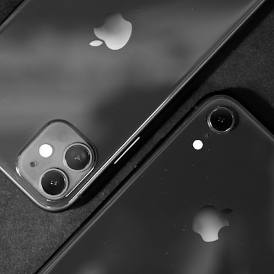 Two smartphones with prominent Apple logos are positioned closely together. One phone features a dual-camera setup, while the other has a single camera. The image is in black and white, creating a sleek and modern aesthetic.