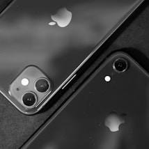 Two smartphones with prominent Apple logos are positioned closely together. One phone features a dual-camera setup, while the other has a single camera. The image is in black and white, creating a sleek and modern aesthetic.