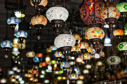 A collection of ornate mosaic lamps with intricate patterns and colorful glass designs suspended from the ceiling. The lamps are of various sizes and shapes, and their vibrant colors stand out against the dimly lit background.