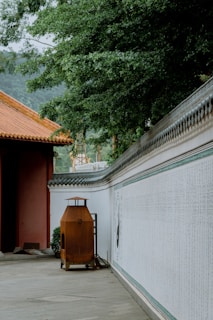 A traditional courtyard scene with a tiled orange roof, a tall rust-colored incense burner, and a long white wall adorned with intricate, vertical calligraphy. Dense green foliage and distant tree-covered hills frame the left side, adding a natural element to the architectural setting.