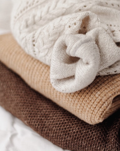 Three stacked knitted sweaters in different textures and earthy tones are displayed, with a creamy white cable-knit sweater on top, a light brown ribbed knit in the middle, and a rich brown chunky knit at the bottom.