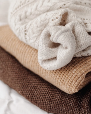 Three stacked knitted sweaters in different textures and earthy tones are displayed, with a creamy white cable-knit sweater on top, a light brown ribbed knit in the middle, and a rich brown chunky knit at the bottom.