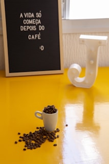 A small white cup filled with coffee beans is placed on a bright yellow surface. Some beans are spilling out of the cup. In the background, there's a letter board with the Portuguese phrase 'A vida só começa depois do café :D' and a decorative white letter 'J'.