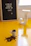 A small white cup filled with coffee beans is placed on a bright yellow surface. Some beans are spilling out of the cup. In the background, there's a letter board with the Portuguese phrase 'A vida só começa depois do café :D' and a decorative white letter 'J'.