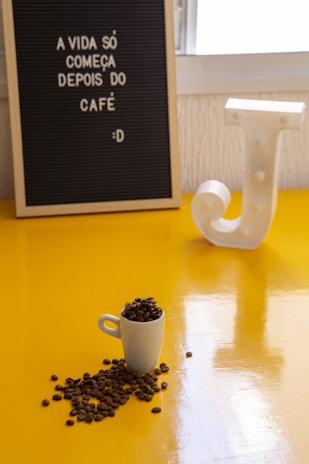 A small white cup filled with coffee beans is placed on a bright yellow surface. Some beans are spilling out of the cup. In the background, there's a letter board with the Portuguese phrase 'A vida só começa depois do café :D' and a decorative white letter 'J'.