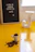 A small white cup filled with coffee beans is placed on a bright yellow surface. Some beans are spilling out of the cup. In the background, there's a letter board with the Portuguese phrase 'A vida só começa depois do café :D' and a decorative white letter 'J'.