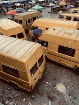 Numerous yellow vans are parked closely together on a muddy street. The ground is wet, suggesting recent rainfall, and there are scattered puddles. Umbrellas are visible, held by people navigating the area. In the background, there is a street market with various colored tents, and vehicles can be seen driving by.