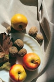 A composition featuring apples and walnuts arranged on a white plate placed on a textured fabric surface. The apples have a vibrant red and yellow hue, and the walnuts are scattered, accompanied by some dry leaves. The setting is bathed in soft, warm sunlight, creating gentle shadows.