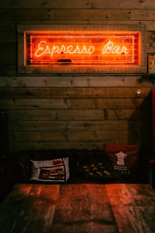 A warmly lit interior featuring a neon sign with the words 'Espresso Bar' mounted on a wooden plank wall. Below the sign, there is a cozy seating area with dark cushioned couches and patterned throw pillows, creating a relaxed atmosphere. A wooden table is in the foreground, slightly out of focus.