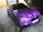 A car adorned with a vibrant galaxy-themed wrap featuring various shades of purple, accented with white spots resembling stars. The vehicle is parked indoors on a concrete surface, with tiles and brickwork visible in the background. Blacked-out windows and sporty black rims enhance its sleek appearance.