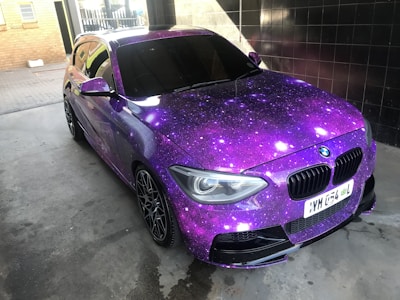 A car adorned with a vibrant galaxy-themed wrap featuring various shades of purple, accented with white spots resembling stars. The vehicle is parked indoors on a concrete surface, with tiles and brickwork visible in the background. Blacked-out windows and sporty black rims enhance its sleek appearance.
