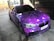 A car adorned with a vibrant galaxy-themed wrap featuring various shades of purple, accented with white spots resembling stars. The vehicle is parked indoors on a concrete surface, with tiles and brickwork visible in the background. Blacked-out windows and sporty black rims enhance its sleek appearance.