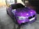 A car adorned with a vibrant galaxy-themed wrap featuring various shades of purple, accented with white spots resembling stars. The vehicle is parked indoors on a concrete surface, with tiles and brickwork visible in the background. Blacked-out windows and sporty black rims enhance its sleek appearance.