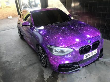 A car adorned with a vibrant galaxy-themed wrap featuring various shades of purple, accented with white spots resembling stars. The vehicle is parked indoors on a concrete surface, with tiles and brickwork visible in the background. Blacked-out windows and sporty black rims enhance its sleek appearance.