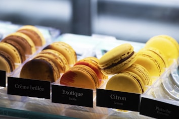 A selection of colorful macarons displayed in a well-lit showcase, each labeled with its flavor such as Crème brûlée, Exotique, and Citron. The macarons are arranged neatly in rows, showcasing a variety of textures and colors.