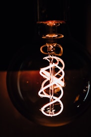An artistic close-up of a glowing filament inside a clear incandescent light bulb, with the filament twisted into a spiral design. The dark background emphasizes the bright, warm orange and yellow hues of the filament.