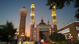 A grand mosque with intricate architecture, featuring two tall minarets adorned with golden lighting. The building is illuminated against the backdrop of a dusky sky, with a prominent green dome and Arabic writing. The surrounding area includes trees, a lamppost, and a few other buildings with lit-up signs.