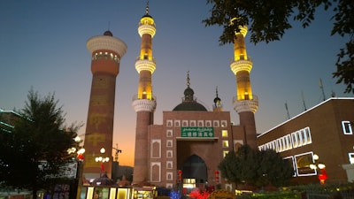 A grand mosque with intricate architecture, featuring two tall minarets adorned with golden lighting. The building is illuminated against the backdrop of a dusky sky, with a prominent green dome and Arabic writing. The surrounding area includes trees, a lamppost, and a few other buildings with lit-up signs.