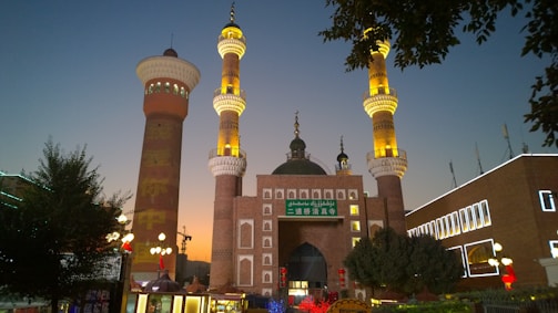 A grand mosque with intricate architecture, featuring two tall minarets adorned with golden lighting. The building is illuminated against the backdrop of a dusky sky, with a prominent green dome and Arabic writing. The surrounding area includes trees, a lamppost, and a few other buildings with lit-up signs.