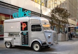 A vintage silver food truck with a large turquoise sign reading 'coffee' is parked on a city street. The truck features a menu on its side and several buildings reflecting in its windows. There are tables with people seated in the background, along with modern city architecture.